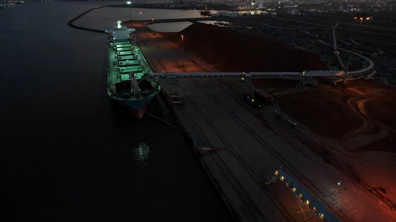 Ein Massengutfrachter wird in der Dämmerung an einem Erzhafen beladen, daneben Halden und Förderbänder.