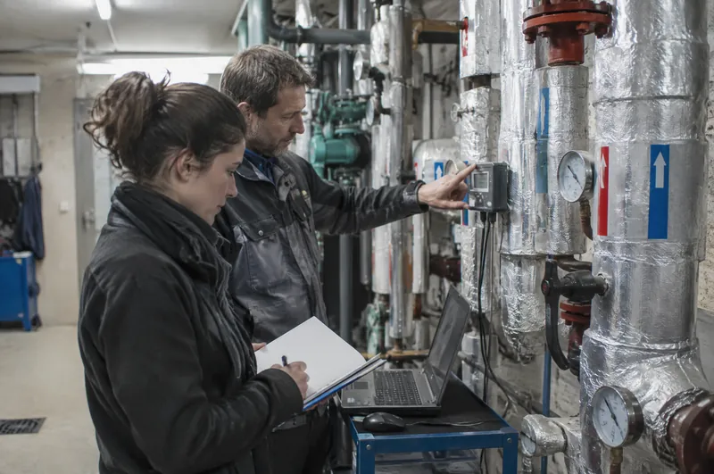 Eine Energiemanagerin und ein Haustechniker lesen im Technikraum einen Wärmezähler ab.