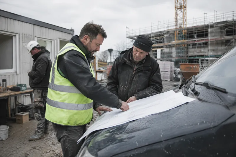 Kurze Baubesprechung am Container: Ein Bauleiter in Warnweste und ein Polier sehen sich Pläne auf der Motorhaube eines Transporters an.