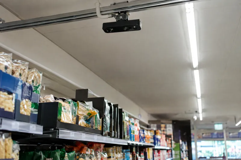 Ein kleiner Tiefensensor an einer Deckenschiene über einem Supermarktgang.
