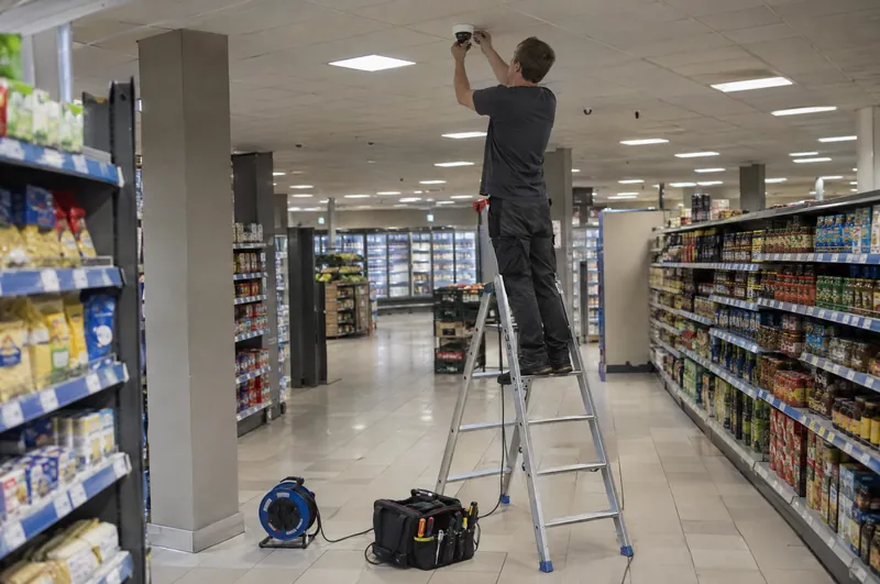 Ein Techniker montiert auf einer Trittleiter eine Kuppelkamera an der Decke eines leeren Supermarkts.